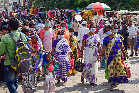 A crowded outdoor market in Hyderabad, India, filled with people walking along a paved path. Several women in the foreground wear colorful traditional sarees and surgical masks, while a man carries a backpack on the left. Clothing stalls and umbrellas line the background under bright daylight.