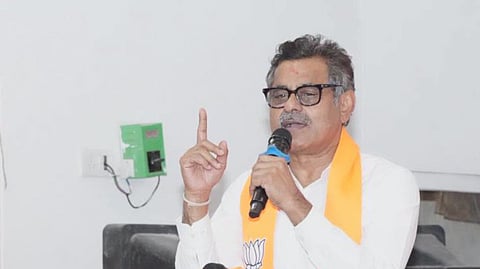 Indian politician Konda Vishweshwar Reddy speaking into a microphone at a podium. He wears glasses, a white shirt, and an orange Bharatiya Janata Party (BJP) stole around his neck, raising his right index finger as he addresses the audience. The podium features a banner displaying the green and saffron colors of the BJP along with its lotus symbol.