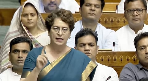Indian politician Priyanka Gandhi Vadra speaking animatedly in Parliament while gesturing with her index finger. She is wearing round spectacles and a dark teal saree with a golden border. Other Members of Parliament are seated in rows behind her, listening in the chamber.