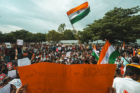 A large crowd of students and young people gathers at Freedom Park in Bengaluru for an education protest. Several Indian flags are raised above the crowd, while protesters hold handwritten placards and banners demanding accountability and access to public education.