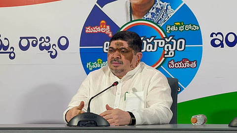 A political leader speaking at a press conference. In the center, Ponnam Prabhakar—an Indian politician dressed in a white traditional kurta—is seated at a table behind a Bosch desktop microphone with his hands gently folded. Behind him is a large backdrop displaying political branding and text written in Telugu script, arranged in a circular design highlighting various welfare schemes (including Maha Lakshmi, Rythu Bharosa, Gruha Jyothi, and Cheyutha). A water bottle is visible on the desk to his left.