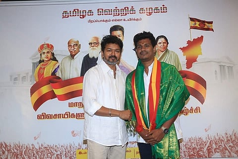 Actor and politician Vijay standing next to another man who is wearing a green shawl and a red-and-yellow sash. They are posing together in front of a political backdrop for TVK (Tamizhaga Vetri Kazhagam). The banner behind them features the TVK party logo, a map of Tamil Nadu, and portraits of historic Tamil leaders and icons including Rani Velu Nachiyar, K. Kamaraj, Periyar E. V. Ramasamy, and Anjalai Ammal.