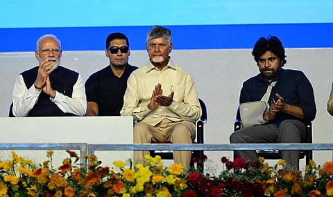 Prime Minister Narendra Modi, Chief Minister N. Chandrababu Naidu, and Deputy Chief Minister Pawan Kalyan sitting together on a stage at a public event, clapping with floral decorations in the foreground.