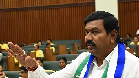 An Indian politician standing at a podium inside a legislative assembly chamber, speaking with his right hand raised in a gesture of emphasis. He is wearing a white shirt and a blue and green party scarf featuring the image of Y.S. Jagan Mohan Reddy and the YSR Congress Party symbol. Microphones are mounted on the wooden podium in front of him, and other assembly members are visible in the background.