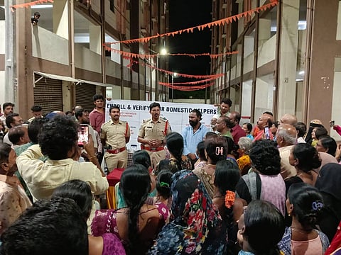 A large crowd gathered in an outdoor courtyard between multi-story residential buildings at night, listening to two police officers in uniform who are speaking in front of a white banner that reads, "ENROLL & VERIFY YOUR DOMESTIC HELPERS."