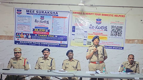 A photo taken inside a hall showing five Telangana police officers in khaki uniforms seated and standing at a long white folding table covered with a black tablecloth. A female police officer stands at the center holding a microphone, speaking to an unseen audience. To her right sit three male police officers, and to her far left sits another male police officer. Behind them hangs a large blue and white banner for "MEE SURAKSHA" (Systematic Enumeration and Verification of Domestic Help Workers) by the Malkajgiri Police Commissionerate, featuring statistics, emergency helpline numbers, a QR code, and instructions for enrolling domestic workers and tenants.