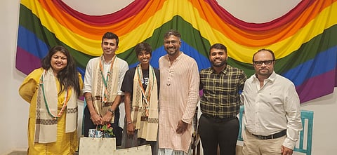 Six people stand in front of a large rainbow Pride flag at a felicitation event in Bengaluru honouring Gay Games 2026 medallists Gowthami Kandhasami (Goaty) and Sumit Pawar. Gowthami and Sumit wear ceremonial shawls and medals, flanked by organisers and guests at 1Shanthiroad Studio/Gallery.