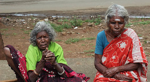 Two Soliga tribal women sit outdoors in Chamarajanagar district, Karnataka. One wears a green blouse with a patterned skirt, while the other wears a red sari and blue blouse.