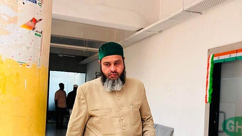 A middle-aged South Asian man with a graying beard, wearing a green prayer cap (topi) and a beige buttoned sherwani coat over white pants, walks down an indoor corridor holding a smartphone. A yellow pillar decorated with Indian flag decals stands in the foreground on the left.
