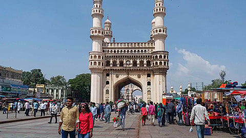 The historic Charminar monument in Hyderabad standing tall under a clear blue sky, surrounded by a bustling street market with pedestrians and vendors.