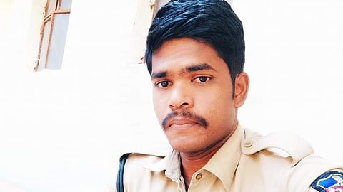 A police officer in a khaki uniform wearing a wrist watch, sitting outdoors near a building wall.