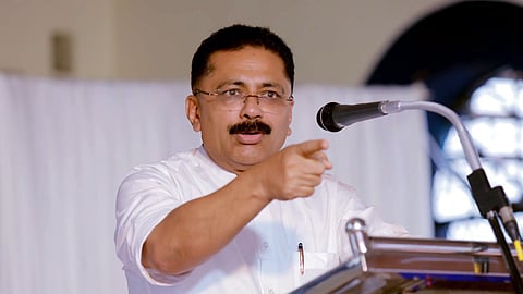 K T Jaleel speaking into a microphone, his hands pointing toward the audience 