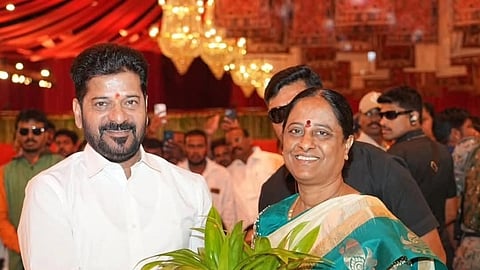 Telangana Chief Minister A. Revanth Reddy, dressed in a crisp white shirt, stands beside Konda Surekha, who is wearing a teal and cream saree. Both are smiling and holding a small potted plant together in a white ceramic pot. The background shows an indoor venue with warm lighting and several guests looking on.