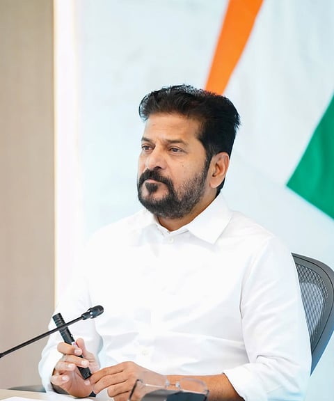 A portrait of Telangana Chief Minister A. Revanth Reddy sitting at a table with a microphone, wearing a crisp white shirt, with an Indian tricolor flag blurred in the background.
