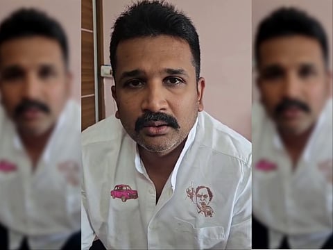 A close-up portrait of Krishank Manne wearing a white button-up shirt featuring small embroidered logos of a pink car on the right chest and a portrait sketch above the left chest pocket. He has short dark hair, a dark mustache, light stubble, and is looking directly into the camera with a neutral expression.   