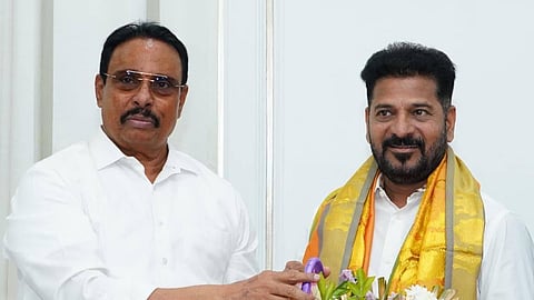 Danam Nagender presenting a large flower arrangement to Revanth Reddy. Danam Nagender, on the left, wears a white shirt and rimless eyeglasses. Revanth Reddy, on the right, wears a white shirt draped with a yellow and orange traditional shawl (kanduva) as he holds the floral basket.   