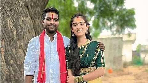 A young Indian couple smiling together outdoors, with the man wearing a white shirt and a red scarf over his shoulders, and the woman wearing a dark green embroidered ethnic attire with traditional jewelry and bangles. Both have red tilak marks on their foreheads.   