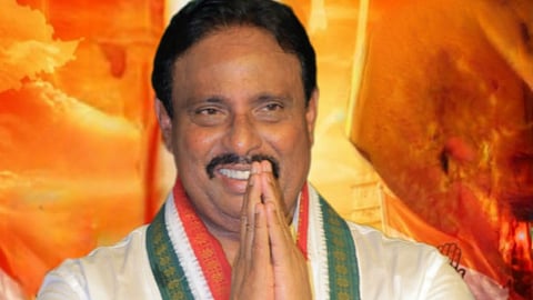 A man with dark hair and a mustache smiling with hands folded together in a traditional greeting ("Namaste"). He is wearing a white shirt and a green, red, and white patterned scarf over his shoulders set against a warm orange-toned background.