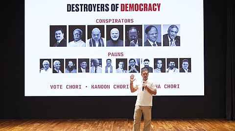 Rahul Gandhi stands on a stage addressing an audience, holding a microphone in one hand and raising his other hand. Behind him, a large presentation screen displays the bold heading "DESTROYERS OF DEMOCRACY". Underneath, two rows of portraits are categorized under red subheadings: the top row is labeled "CONSPIRATORS" and features seven prominent figures including Gautam Adani, Mohan Bhagwat, Amit Shah, and Narendra Modi; the bottom row is labeled "PAWNS" and features eleven figures. At the bottom of the screen, text reads "VOTE CHORI • KANOON CHORI • [...] CHORI".   