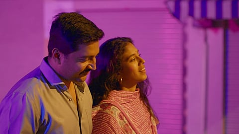 A still from 'Heggana Muddu'. A man and a woman walking side by side on a street. Lighting is purple