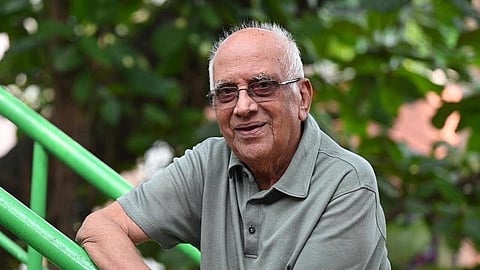 An elderly South Indian man with short white hair and eyeglasses, smiling gently while leaning against a bright green metal staircase handrail outdoors with foliage in the background.   