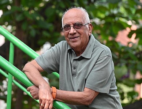 An elderly South Indian man with short white hair and eyeglasses, smiling gently while leaning against a bright green metal staircase handrail outdoors with foliage in the background.   