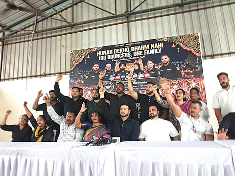 A group of bouncers and organizers seated behind a table at a press conference, many raising their hands or fists in unity. Behind them stands a large banner displaying the headline "HUNAR DEKHO, DHARM NAHI" and "100 BOUNCERS, ONE FAMILY" along with portraits of male and female bouncers from diverse religious backgrounds.