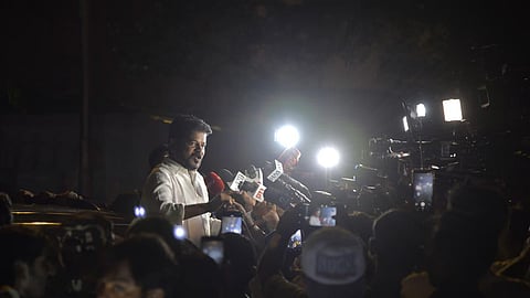 Telangana Chief Minister Revanth Reddy speaking to news media and reporters at night surrounded by camera lights and microphones.   