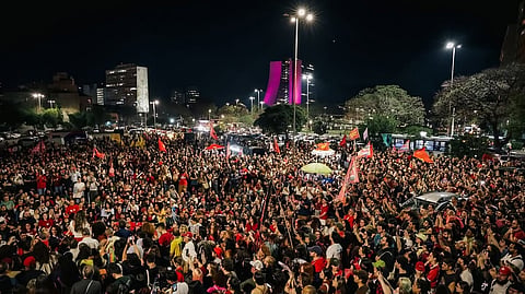 A large crowd of protesters fills a city street at night in Rio de Janeiro, Brazil, waving red flags and banners during a demonstration. Buildings, streetlights, trees, and vehicles are visible in the background.