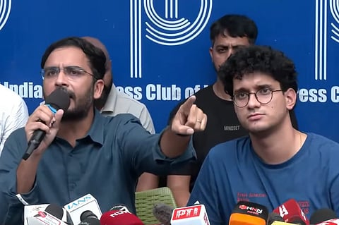 CJP leaders Saurav Das and Ashutosh Ranka speaking at a press conference at the Press Club of India. On the left, Ashutosh wears glasses and a dark shirt, holding a microphone in his right hand and gesturing with his index finger. On the right, Saurav wears round glasses and a blue t-shirt, looking toward the camera. Multiple news microphones (including IANS, PTI, and NDTV) sit on the table in front of them.   