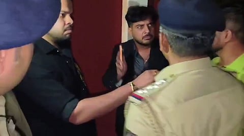 A man in a black shirt reaches out toward a police officer in a khaki uniform, while another man in a dark blazer standing behind him holds up a mobile phone with a tense expression.   