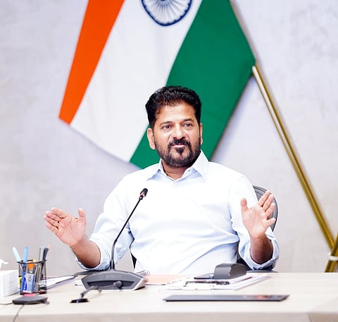 A medium close-up shot of A. Revanth Reddy, Chief Minister of Telangana, seated at a desk speaking with open hands in a gesturing pose. He is wearing a light blue button-down shirt, and the Indian national flag is positioned in the background behind him. A conference microphone, desk organizer, and papers are visible on the table in front of him.
