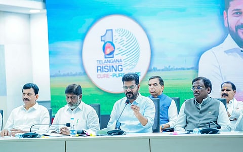 Chief Minister of Telangana, A. Revanth Reddy, sitting at a conference table with a microphone, gesturing while speaking during a meeting. He is seated alongside other government officials and ministers. Behind them is a large screen displaying the logo "TELANGANA RISING CURE-PURE-SECURE" with a landscape background.   