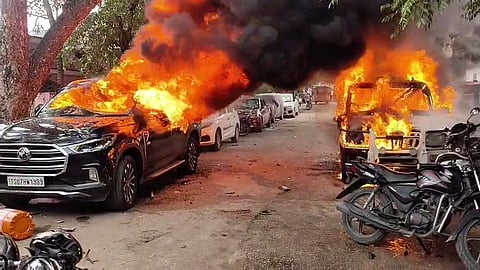 Two vehicles, including a black SUV on the left, engulfed in large orange flames and thick black smoke on a street lined with parked cars and motorcycles.   