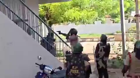 A security officer wearing tactical gear pointing a handgun upward near a white outdoor staircase. Other security personnel wearing helmets and protective vests with "AP POLICE" printed on the back stand nearby in an outdoor area surrounded by greenery and a light-colored boundary wall.   
