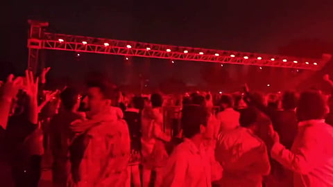 A large crowd gathered at a night event illuminated by intense red stage lighting overhead. A horizontal overhead truss with a row of bright red lights spans across the dark background above the audience.   