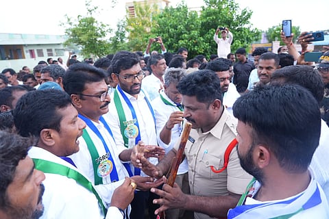 A police officer in uniform holding a lathi stands surrounded by a group of men wearing YSRCP political sashes featuring party colors and symbols during a outdoor gathering.