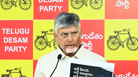 N. Chandrababu Naidu holding up a printed placard featuring a photo and Telugu text during a press conference against a yellow and red Telugu Desam Party backdrop.