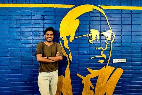 Mathur Sathya standing with his hands folded behind the blue and yellow background of Ambedkar