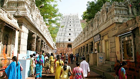 People walk towards a large, intricately carved temple in Tirumala in an outdoor setting, surrounded by trees and detailed stone architecture