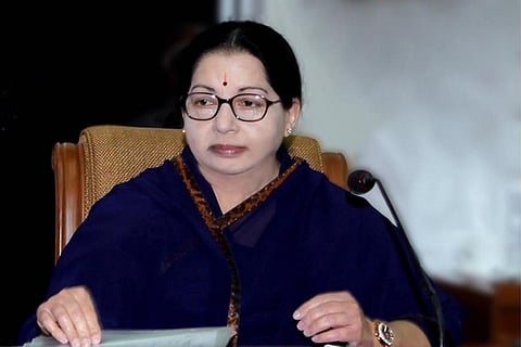 Late Tamil Nadu Chief Minister J Jayajalithaa is seated at a mic, wearinf a dark blue saree, glasses and a pottu. The background behind her is black and white. 