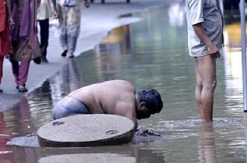 A man waist deep in a 'man hole' on a water logged road. Pedestrians walking on the dry part of the road. 
