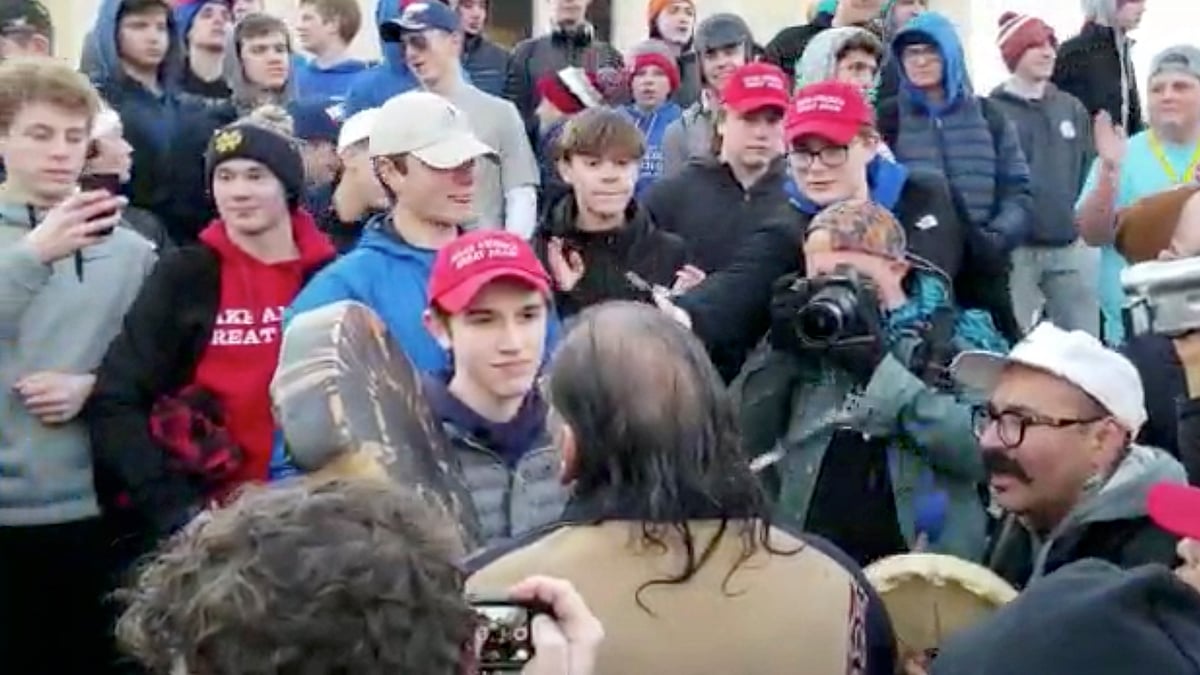 In this Friday, 18 January, image made from video provided by the Survival Media Agency, a teenager wearing a “Make America Great Again” hat, center left, stands in front of an elderly Native American singing and playing a drum in Washington. 