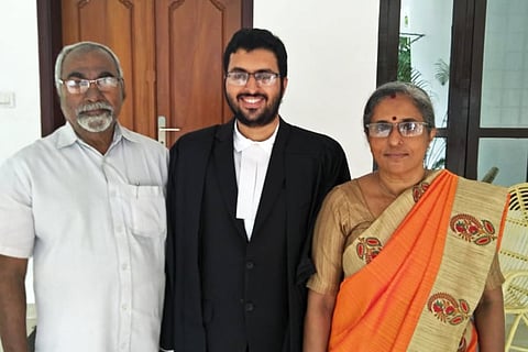 A young man in lawyer's coat stands between an older man dressed in white and a woman in an orange saree. 