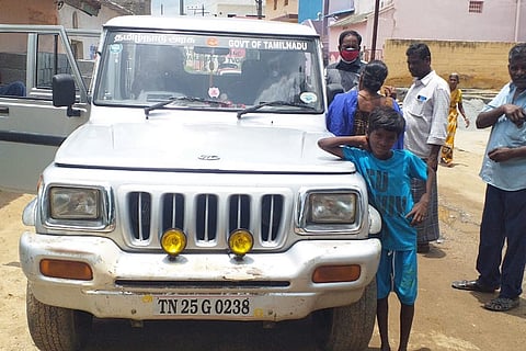 A young boy wearing blue shirt and shots standing close to Tax department white car