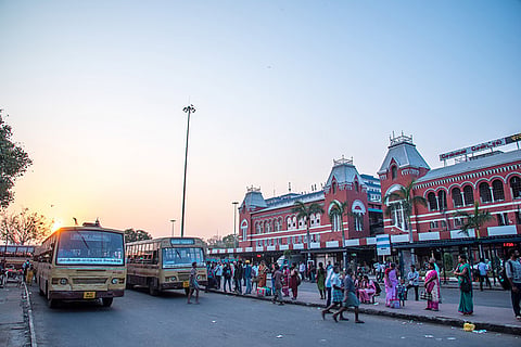 A photograph of the busy street outside the Chennai Central Railway Station in India, which features striking red brick Victorian-Gothic architecture with a clock tower and arched windows. In the foreground, two yellow MTC (Metropolitan Transport Corporation) buses are parked on the left. A large number of pedestrians are visible, some walking on the pavement and others near the buses. The scene is illuminated by the warm, golden light of the setting or rising sun visible on the left side of the frame.