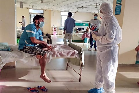 A man sitting on a bed in a hospital wearing a mask, talking to a doctor wearing a PPE suit