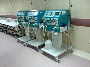 A row of dialysis machines is lined up inside a hospital ward, each connected with tubes and control panels. The machines are placed beside patient beds, indicating a clinical setting for kidney treatment.