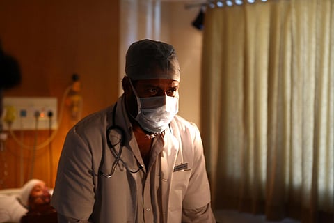 A doctor wears a mask and surgical cap with a stethoscope around his coat 