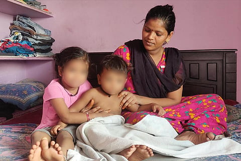 Sathya in a pink nightie wearing a brown shawl across her is sitting on a bed with her two children Sadhana and Eshanth. Eshanth is not wearing a shirt, Sadhana is in a pink dress